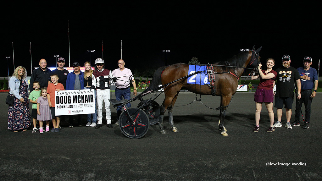 Doug McNair next to Stable Genius in the winner's circle with family holding up a placard reading "Congratulations Doug McNair, $100 million in career earnings"