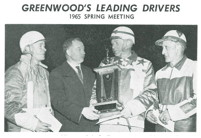 Three drivers in colours stand with a man in a suit, who presents a trophy to the man second from right