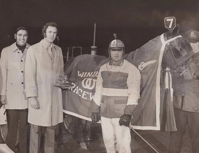 A driver stands in front of a horse in a cooler with two men in coats to the side