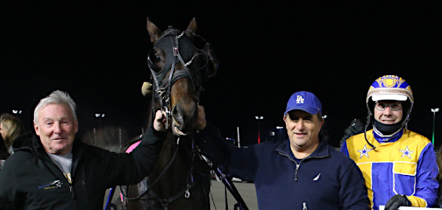 Two men hold a horse's head, with a man in driving colours to their right