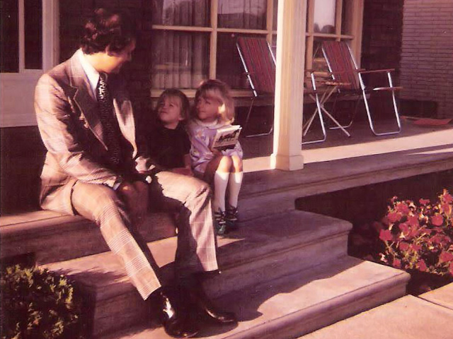 A man in a suit sits on a porch with two young girls