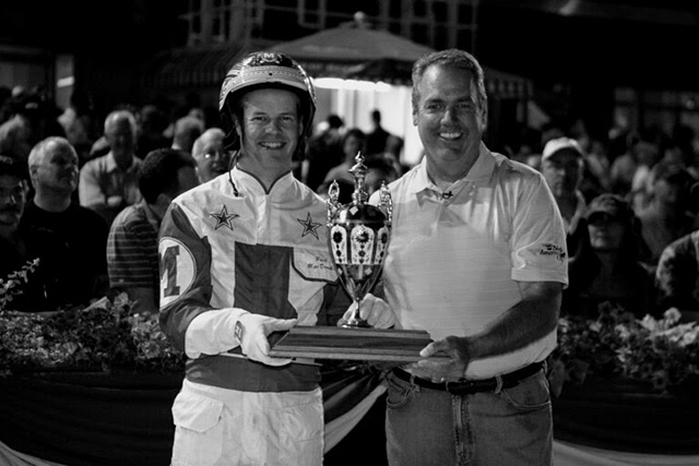 A man in driving colours and a man in a polo shirt hold the North America Cup trophy together