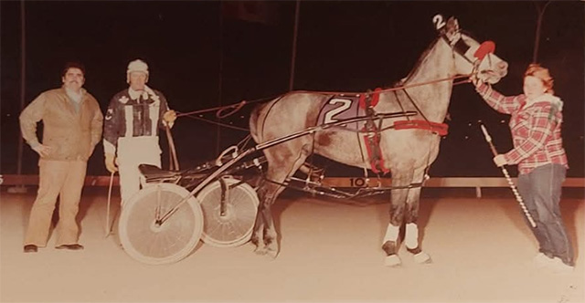 A grey horse in a winner's circle being held by a groom and a man in driving colours