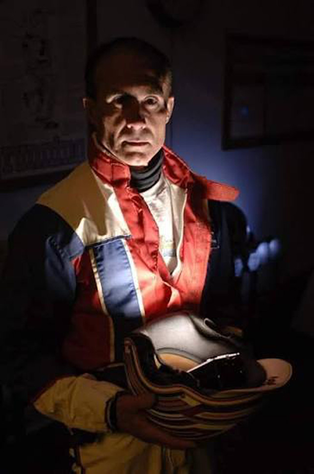 A man in red, white and blue driving colours in a dark room, lit from below