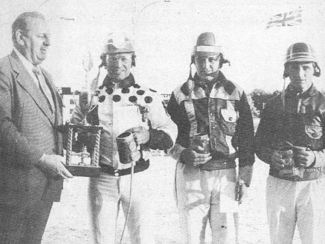 A man in a suit presents a driver with a trophy, with two other drivers holding tankards