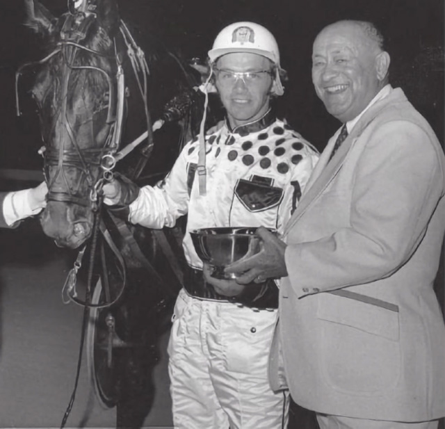 A driver holding a horse holds a bowl trophy with a man in a suit