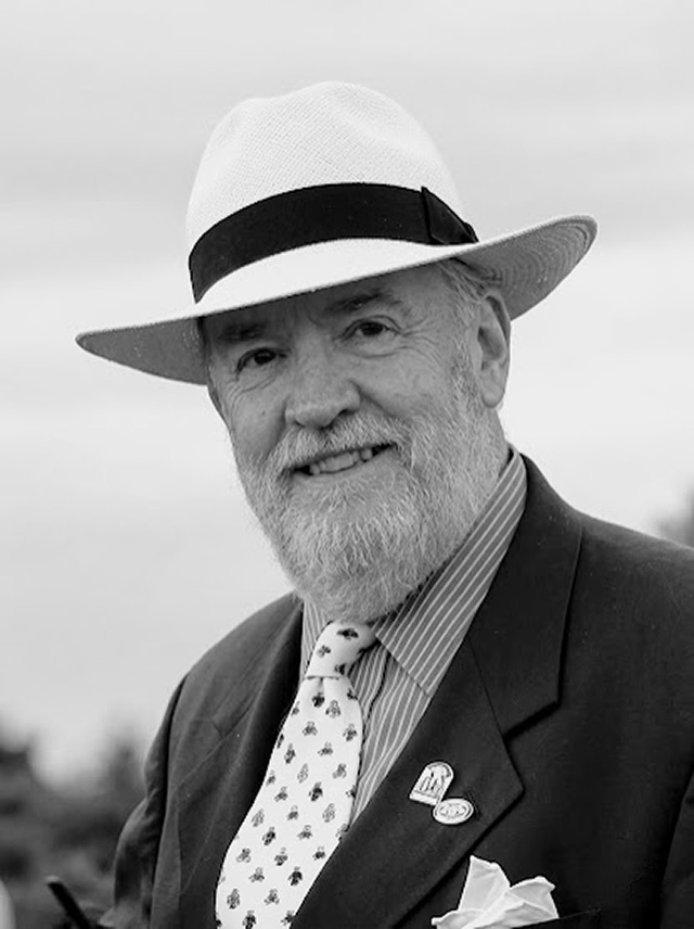 A bearded man in a striped shirt with a tie with racing colours on it, a dark jacket and white panama hat.